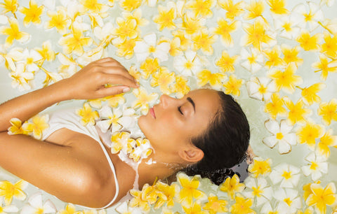 Woman relaxing in water surrounded by yellow and white flowers, eyes closed, serene expression.